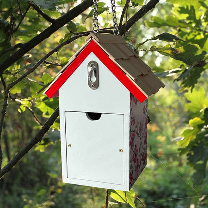 A small house-shaped birdhouse hanging from a chain in a tree. The birdhouse has a white front door, a red roof trim, a brown shingled roof, and floral-patterned sides, set against a blurred green leafy background.