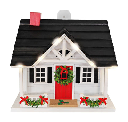 A decorative bird house with a black shingled roof, white walls, a bright red front door and chimney, green wreaths with red bows, garlands, and warm white string lights on a white background.