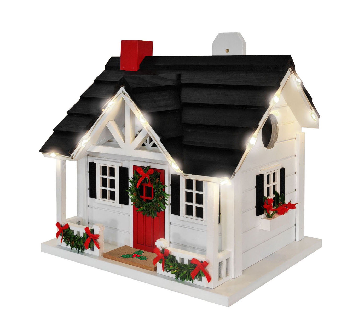 A decorative Christmas birdhouse with a white exterior, black shingled roof, red front door and chimney, green wreath, porch railings with red bows, and warm white string lights along the roofline and windows, shown on a plain white background.