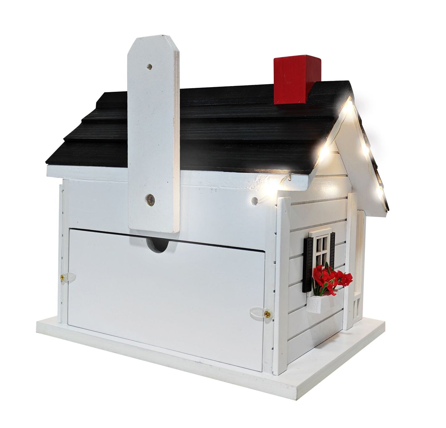 A white wooden birdhouse-style mailbox with a black shingled roof, red chimney, warm white string lights, and a small red flower arrangement on the front.