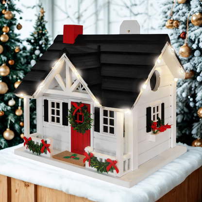 A decorative Christmas birdhouse with a white exterior, black roof, red door and chimney, green wreath and garlands, and warm white string lights, displayed on a snowy surface with Christmas trees in the background.