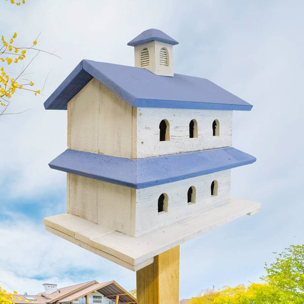 A decorative two-story birdhouse with a cream-colored wooden body and blue-painted roof and eaves, mounted on a wooden post against a cloudy sky background.