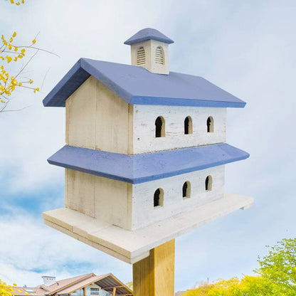 A decorative two-story birdhouse with a cream-colored wooden body and blue-painted roof and eaves, mounted on a wooden post against a cloudy sky background.
