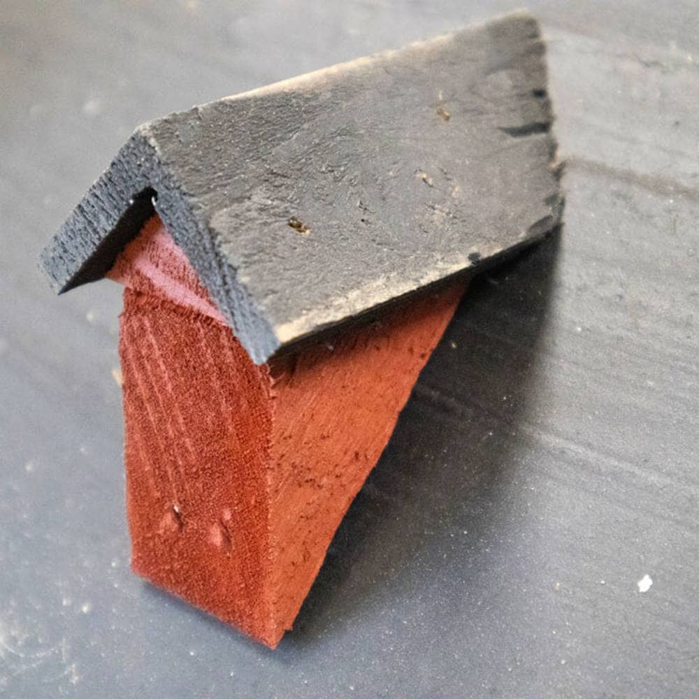 A small handmade miniature house figurine with a dark charcoal gray roof and a reddish-brown wooden body, set against a dark gray textured background.