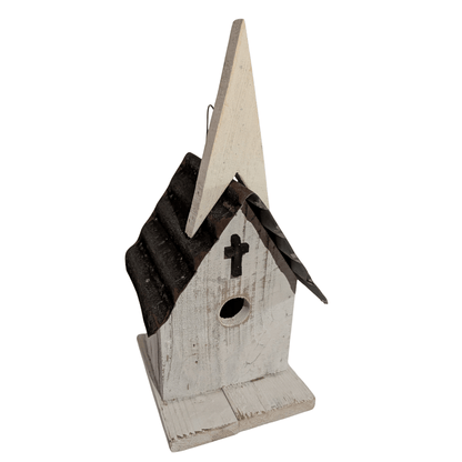 A rustic church-shaped birdhouse with a distressed white painted wood body, a dark brown corrugated metal roof, and a small black cross on the front.