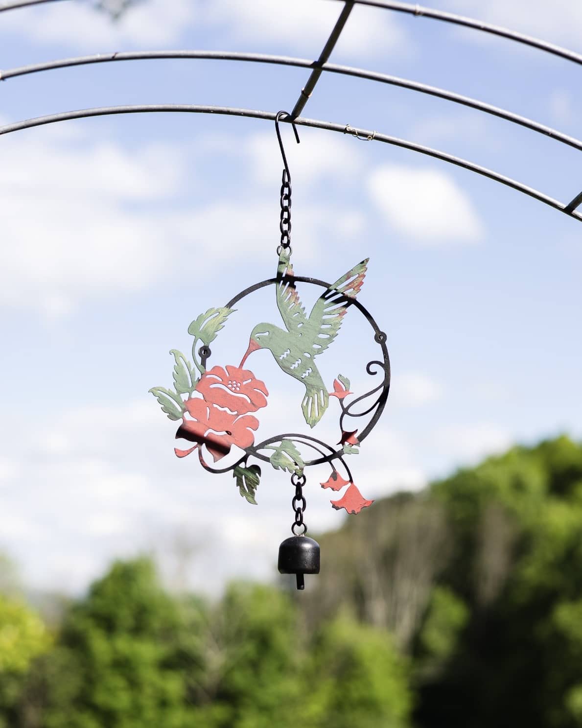 Metal circular garden ornament with green hummingbird, red rose, and trumpet flowers, hanging with a small black bell
