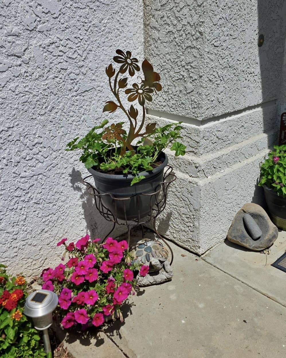 Rusty metal daisy flower garden stake displayed in a dark pot with green plants, surrounded by pink petunias outdoors
