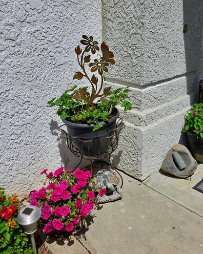 Rusty metal daisy flower garden stake displayed in a dark pot with green plants, surrounded by pink petunias outdoors