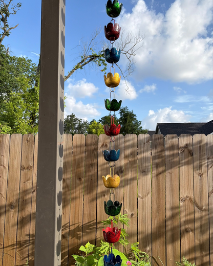 A vertical hanging rain chain of colorful flower-shaped garden ornaments in red, green, blue, and yellow, displayed outdoors against a wooden fence and blue sky.