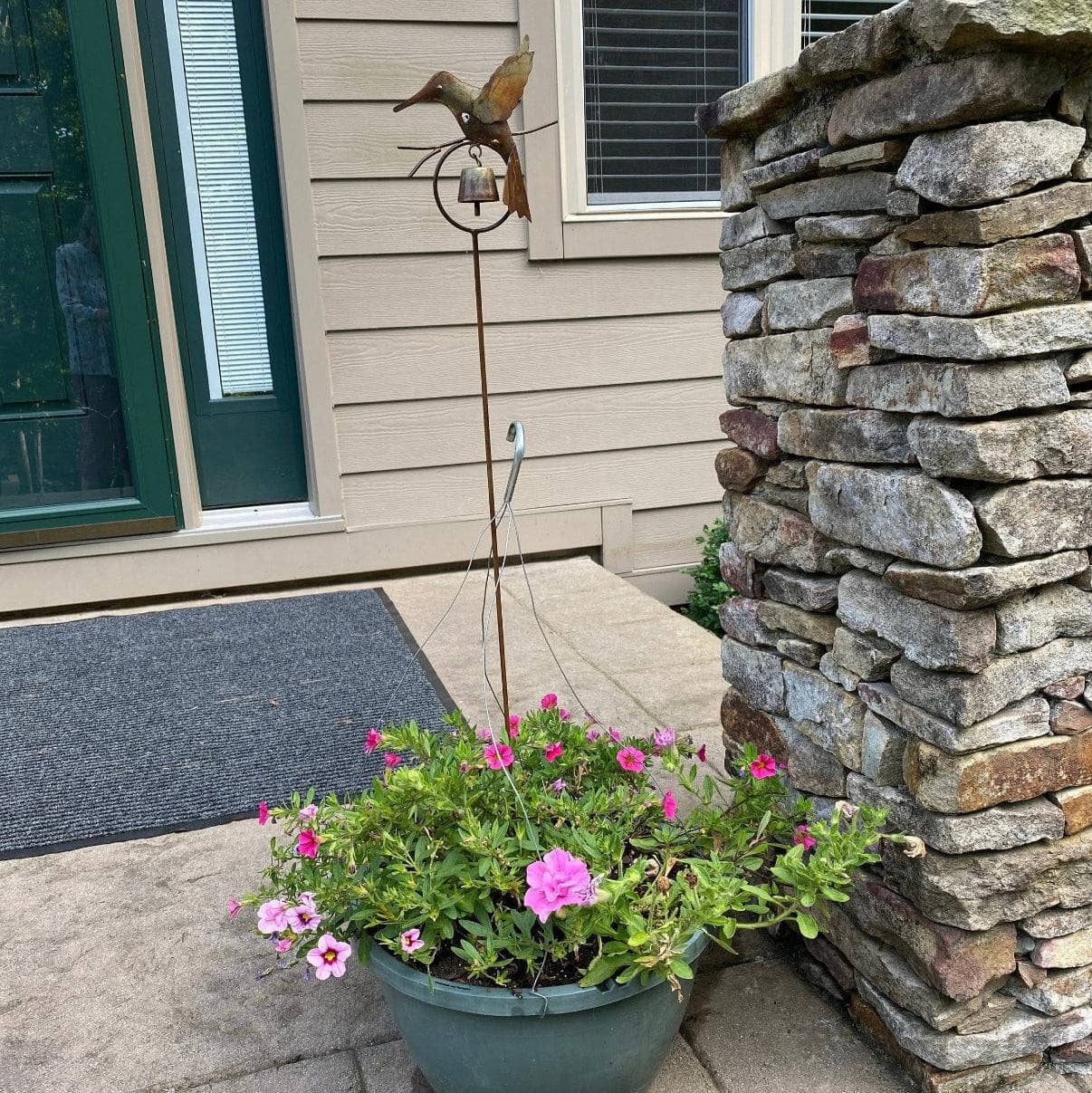 Rusty metal hummingbird garden stake with small bell, placed in a pink flowering potted plant near a stone pillar entryway