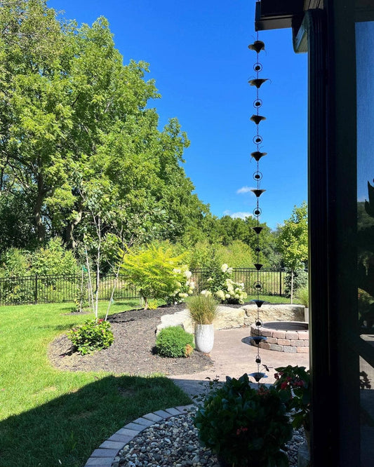 Dark metal rain chain with cup-shaped lily flower links hanging from a rooftop gutter over a landscaped backyard patio
