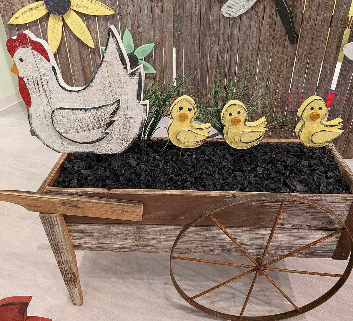 Rustic farm-themed decorative display featuring a large distressed white hen cutout with a red comb, three yellow ducklings, black charcoal-like filler, and a weathered wooden wheel planter against a wood plank backdrop.