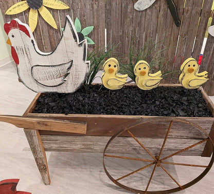 Rustic farm-themed decorative display featuring a large distressed white hen cutout with a red comb, three yellow ducklings, black charcoal-like filler, and a weathered wooden wheel planter against a wood plank backdrop.