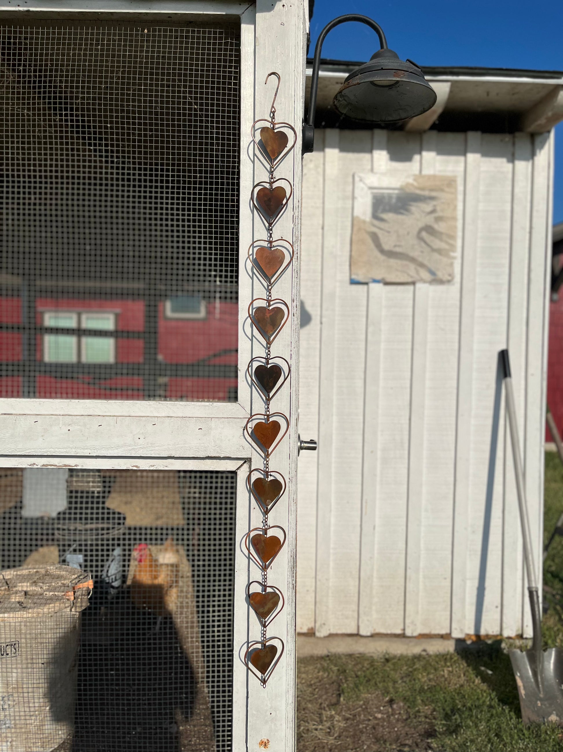 Copper heart-shaped garden ornament hanging from roofline beside gray shutter and window with lace curtains