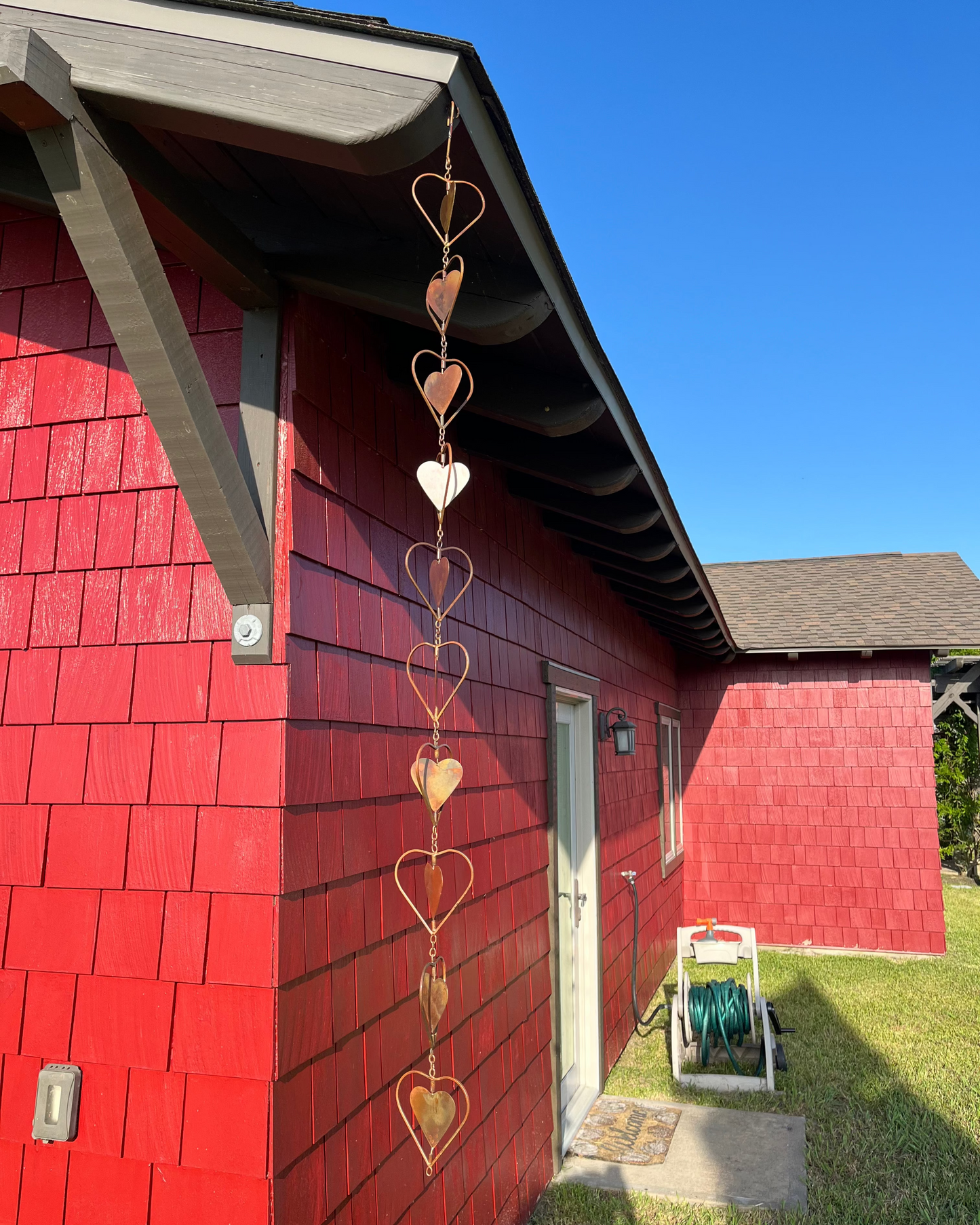 Copper heart-shaped garden ornament hanging from roofline beside gray shutter and window with lace curtains