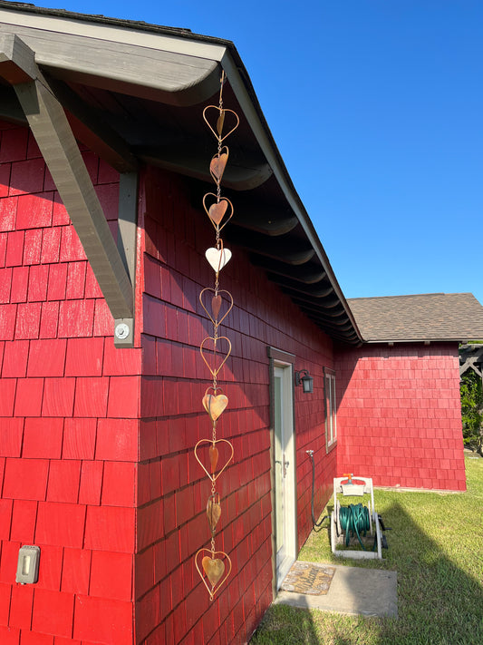 Copper heart-shaped garden ornament hanging from roofline beside gray shutter and window with lace curtains