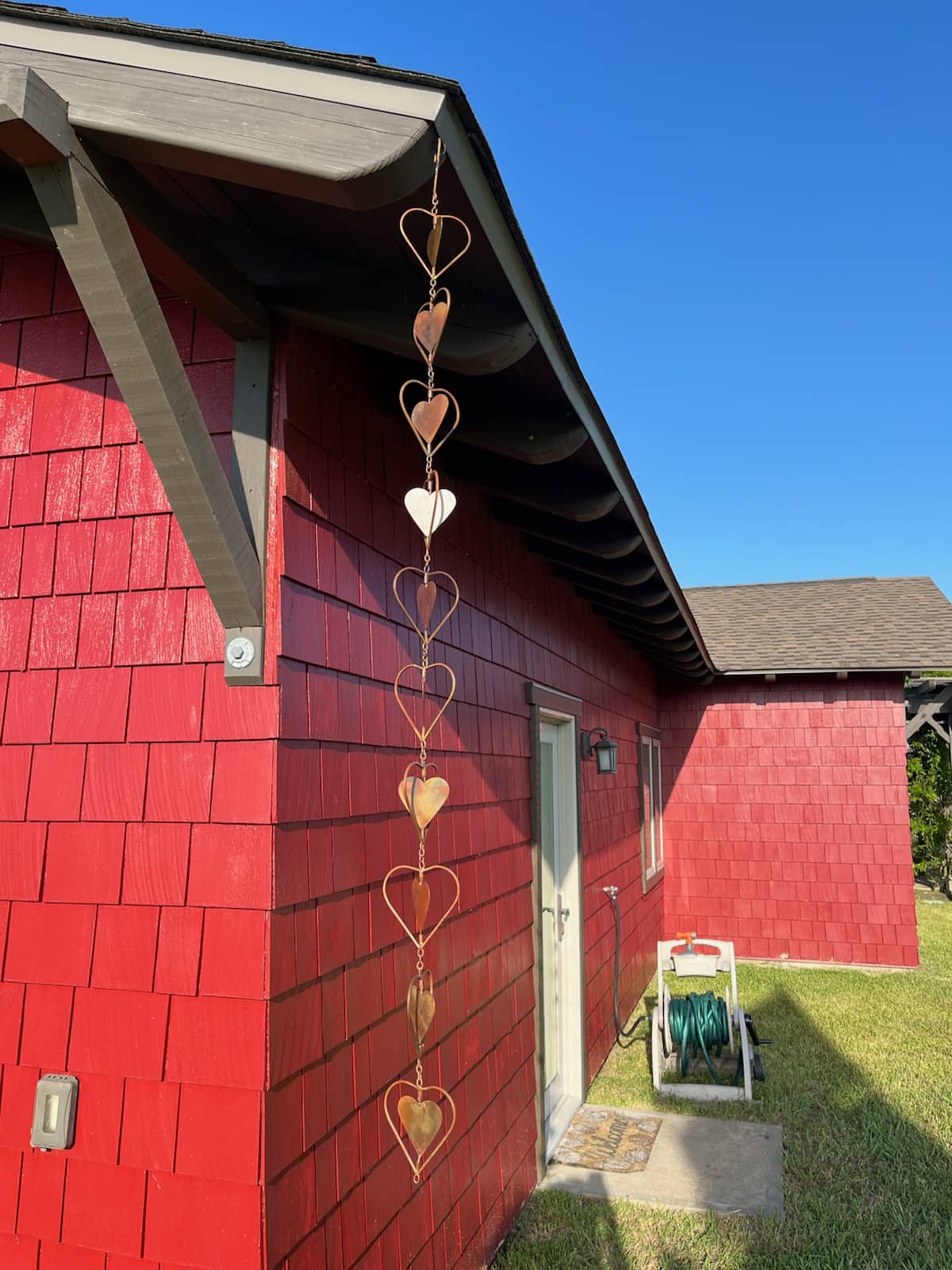 Copper heart-shaped garden ornament hanging from roofline beside gray shutter and window with lace curtains