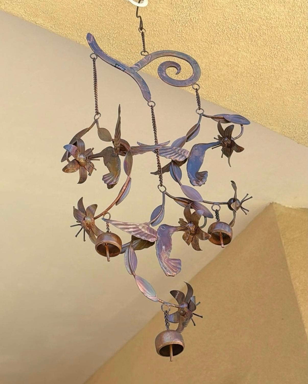 A rustic metal wind chime featuring birds, flowers, and hanging bells, suspended from a textured ceiling in a beige room.