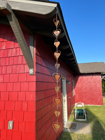 Copper heart-shaped garden ornament hanging from roofline beside gray shutter and window with lace curtains