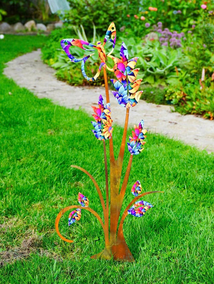 A colorful metal garden sculpture shaped like stylized flowers or leaves, mounted on a grassy lawn with a garden path and plants in the background.