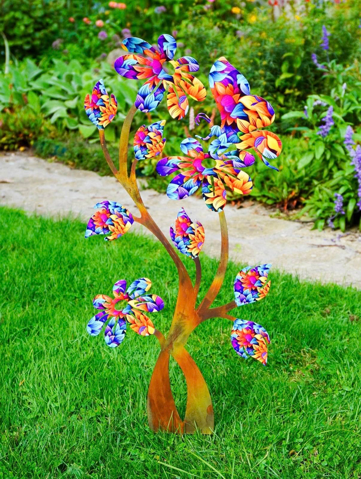A colorful metal garden daisy sculpture with a rusted orange-brown trunk and vibrant, multicolored leaf-like branches, displayed on a green lawn in a garden setting.