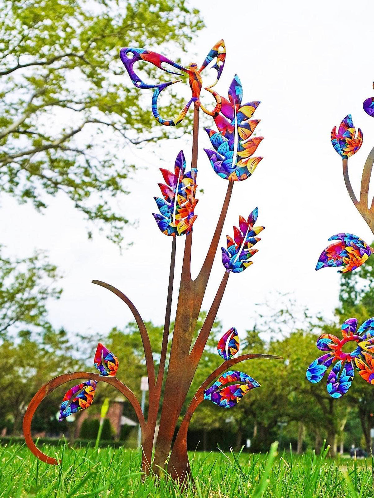Colorful metal garden sculpture of stylized flowers and leaves on a green lawn with trees in the background.
