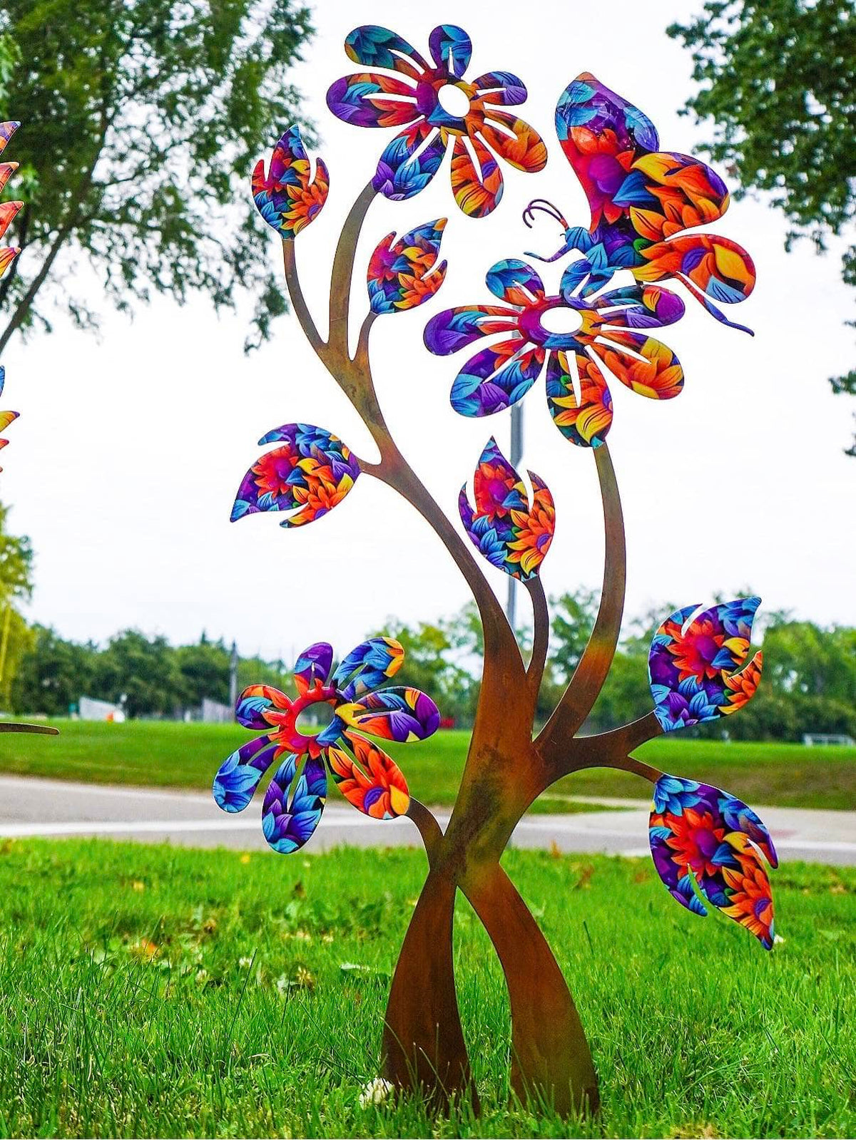 A colorful metal garden sculpture shaped like a tree with flowers and a butterfly, displayed outdoors on a grassy lawn.