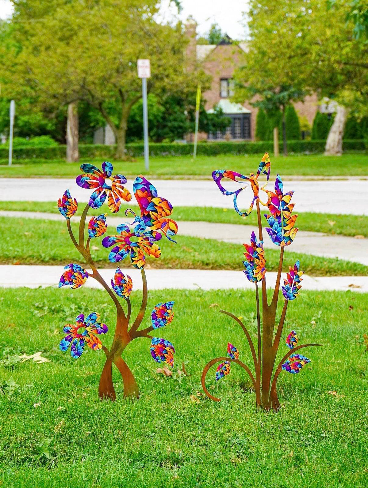 Two colorful metal garden sculptures shaped like stylized flowers or plants, placed on a green lawn in front of a paved path and trees.