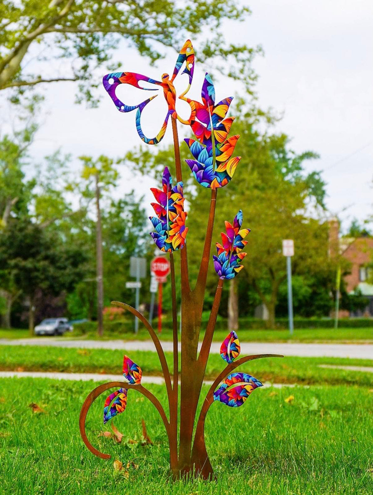 A colorful metal garden sculpture of stylized flowers and leaves on a grassy lawn, with trees and a street in the background.