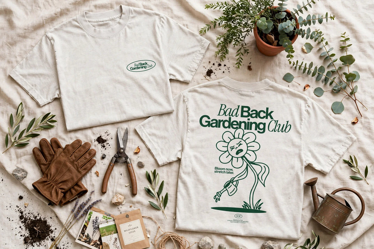 Two white graphic T-shirts arranged on a beige fabric background with gardening props including gloves, shears, leaves, a watering can, and pots. The left shirt shows a small oval chest logo, and the right shirt shows a large back graphic reading 'Bad Back Gardening Club' with a flower illustration.