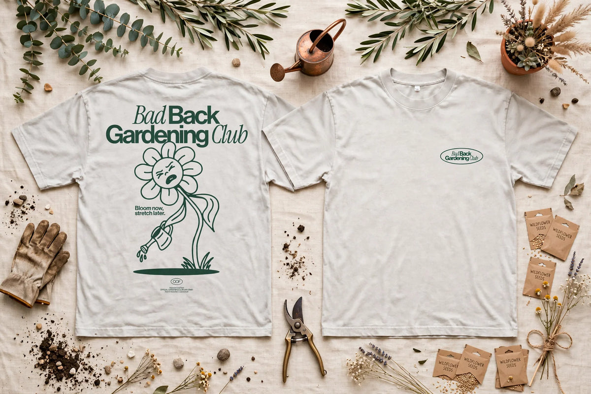 Two white graphic T-shirts laid flat on a light fabric background with gardening props. The left shirt shows the back with the text 'Bad Back Gardening Club' and a large flower illustration; the right shirt shows the front with a small oval 'Bad Back Gardening Club' logo on the left chest.