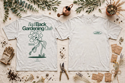 Two white graphic T-shirts laid flat on a light fabric background with gardening props. The left shirt shows the back with the text 'Bad Back Gardening Club' and a large flower illustration; the right shirt shows the front with a small oval 'Bad Back Gardening Club' logo on the left chest.