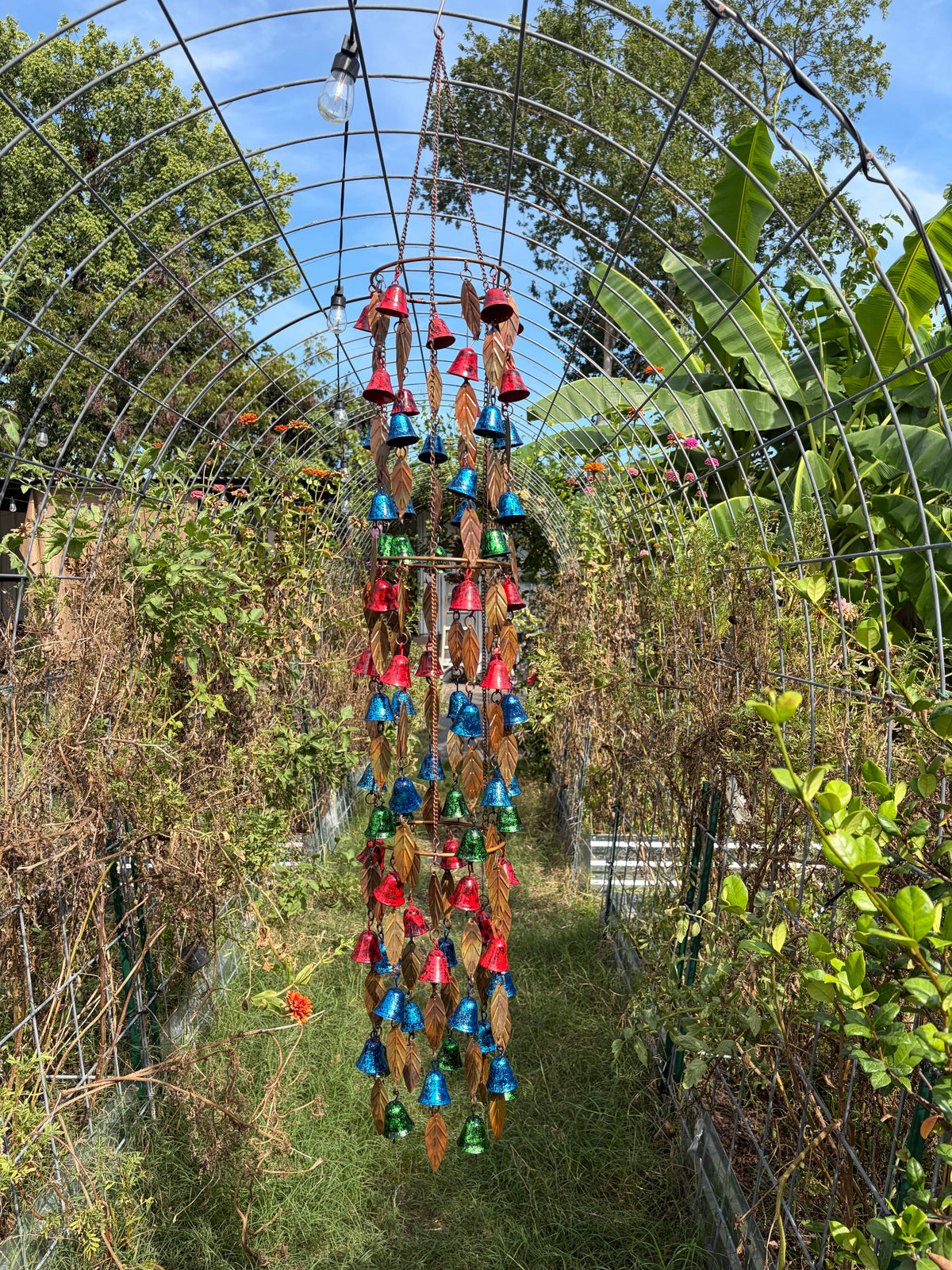 A garden wind chime hanging from a metal trellis arch, featuring colorful bells in red, blue, green, and gold on a brown leaf-shaped frame, set against a lush garden background.