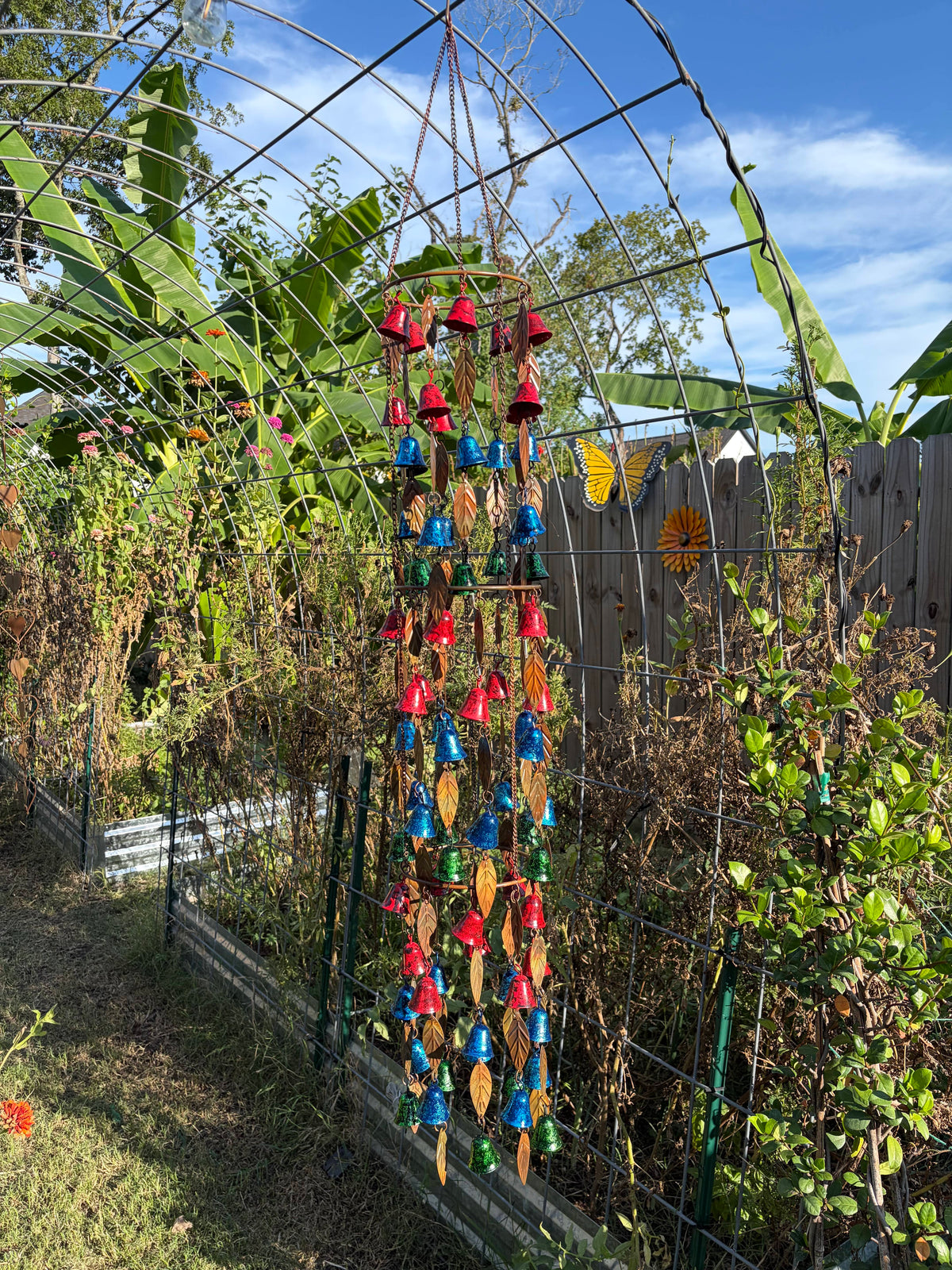 A colorful hanging wind chime garden ornament with red, blue, green, and yellow bells and leaf-shaped pieces, suspended in a backyard garden setting under a metal trellis.