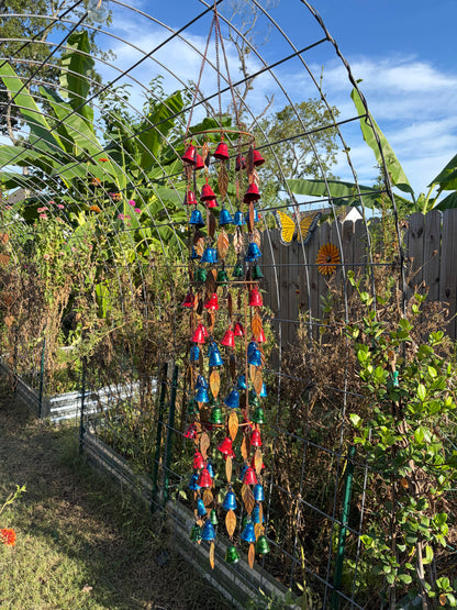 A colorful hanging wind chime garden ornament with red, blue, green, and yellow bells and leaf-shaped pieces, suspended in a backyard garden setting under a metal trellis.