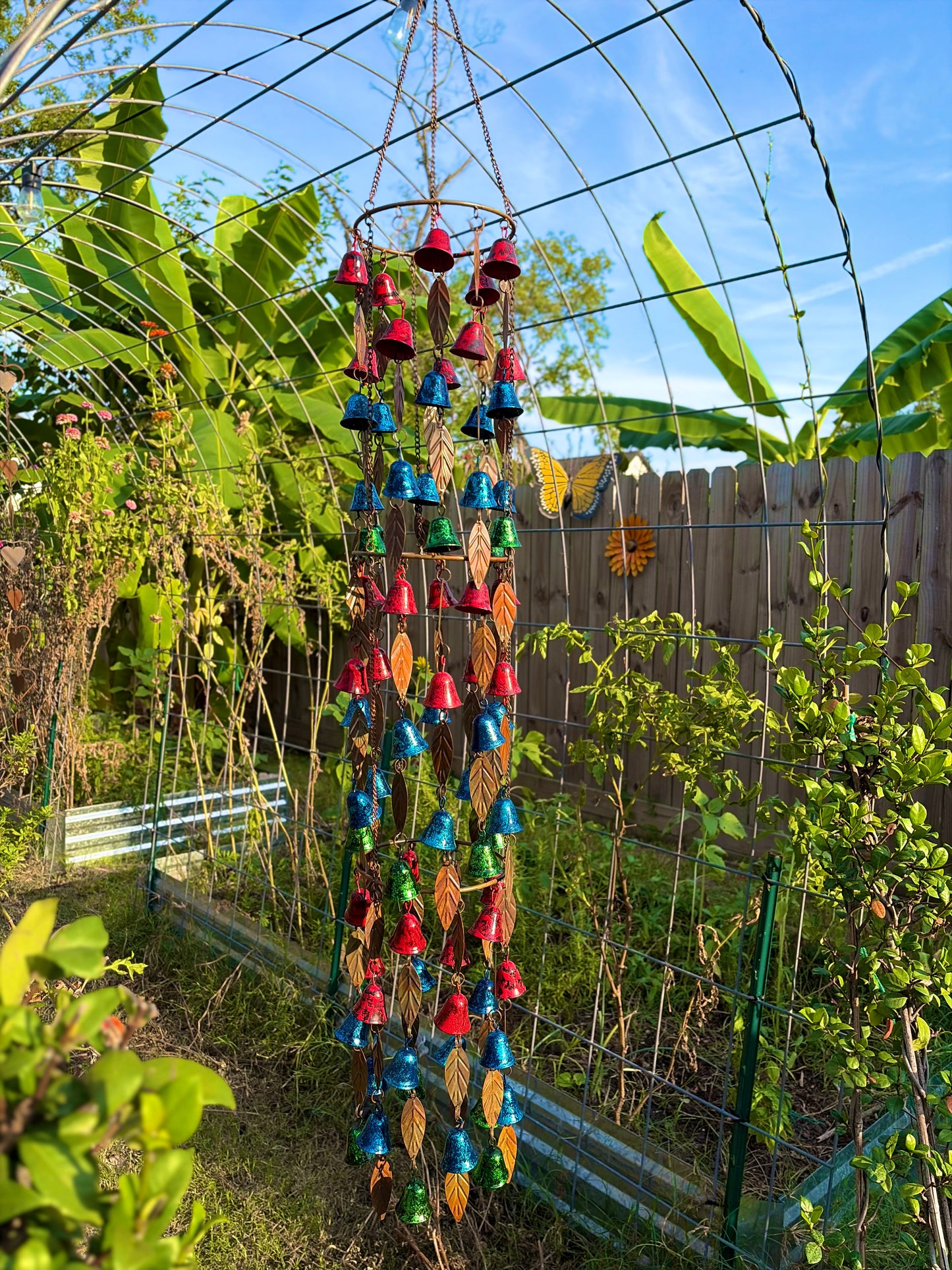A colorful hanging wind chime garden ornament with red, blue, green, and brown leaf-shaped bells suspended from a metal hoop, photographed outdoors in a garden under a greenhouse arch.