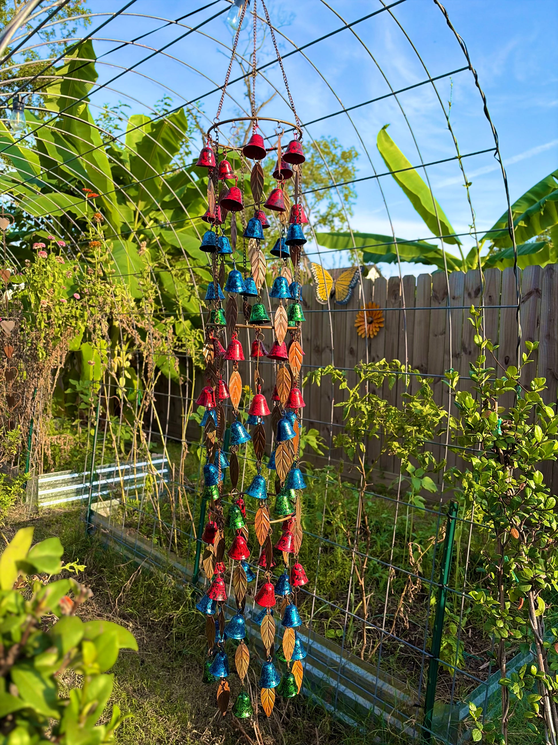 A colorful hanging wind chime garden ornament with red, blue, green, and brown leaf-shaped bells suspended from a metal hoop, photographed outdoors in a garden under a greenhouse arch.