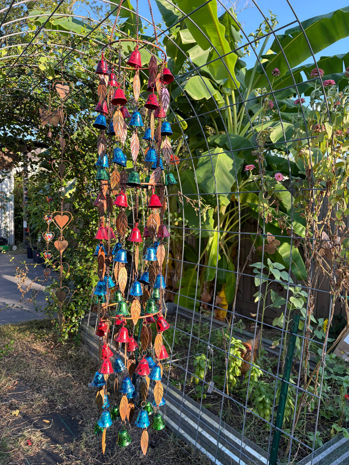 A hanging garden wind chime with colorful metallic bells and dried leaves, displayed outdoors on a wire trellis in a garden with banana leaves and plants in the background.
