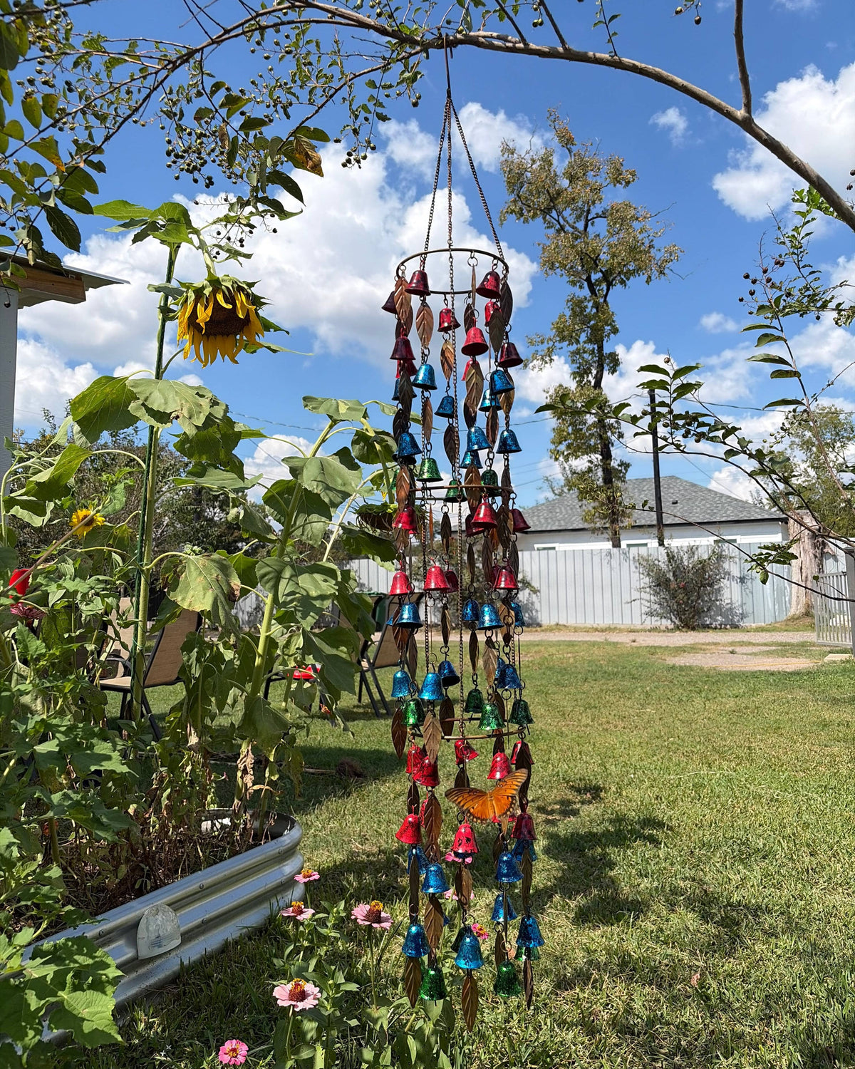 A colorful wind chime garden hanging suspended from a tree branch in a backyard garden. The wind chime features multiple tiers with blue, red, and gold bells, small wind chimes, and butterfly decorations.