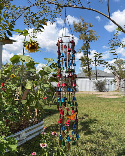 A colorful wind chime garden hanging suspended from a tree branch in a backyard garden. The wind chime features multiple tiers with blue, red, and gold bells, small wind chimes, and butterfly decorations.