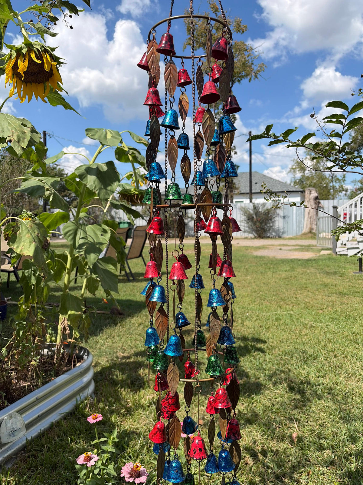A metal wind chime garden ornament hanging outdoors on a grassy lawn under a blue sky with clouds. The chime features multiple tiers of small bells in red, blue, green, and silver, interspersed with brown leaf-shaped ornaments.