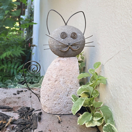 Stone and metal wire cat garden statue with spiral eyes, whiskers, curled tail, sitting outdoors near green plants