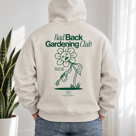 A person wearing a light heather gray hoodie from the back, featuring a dark green graphic of a sad flower and the text 'Bad Back Gardening Club'. The person is also wearing blue jeans, standing in a bright indoor setting with a potted snake plant on the left.