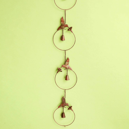 Copper metal wind chime with hummingbird figures, circular rings, and hanging brass bells against a white brick wall