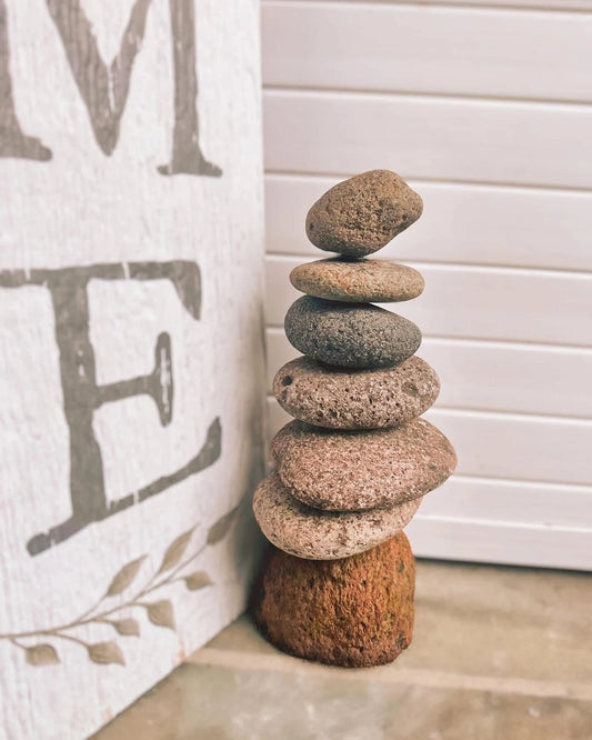 Seven stacked river stones forming a cairn tower on a concrete surface beside a rustic home sign