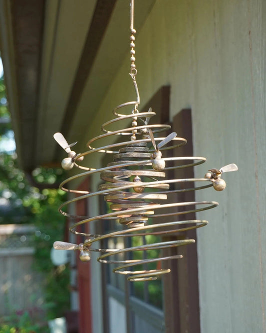 Hanging metal spiral wind spinner with bee figures attached, aged bronze finish, displayed outdoors on a porch