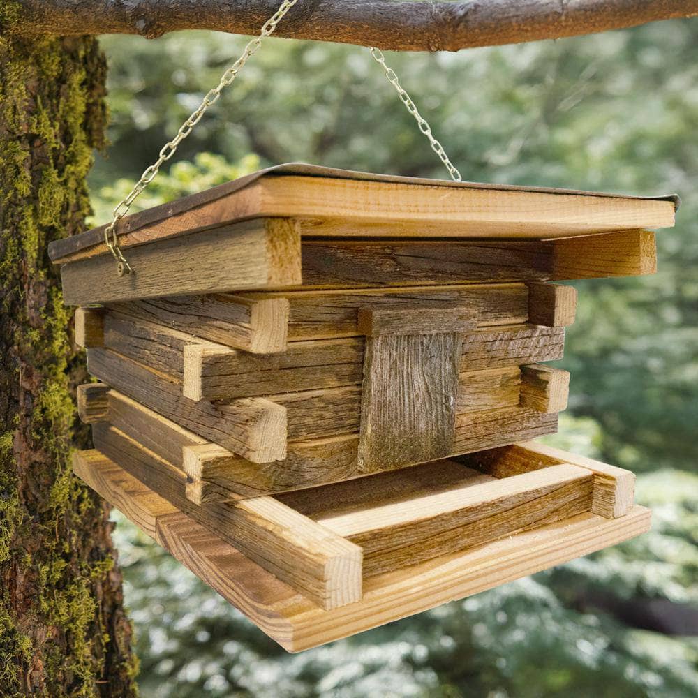 A rustic wooden bird feeder hanging from a metal chain attached to a tree branch outdoors, with blurred green foliage in the background.