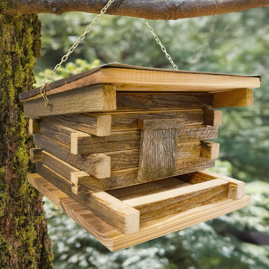 A rustic wooden bird feeder hanging from a metal chain attached to a tree branch outdoors, with blurred green foliage in the background.