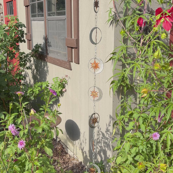 A hanging metal garden star and moon rain chain ornament set displayed outdoors on the exterior wall of a house, surrounded by green plants and flowers.