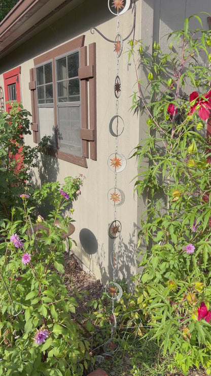 A hanging metal garden star and moon rain chain ornament set displayed outdoors on the exterior wall of a house, surrounded by green plants and flowers.